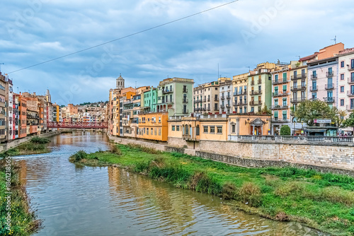 Picturesque view of colorful houses along the Onyar River with the red Eiffel Bridge in historic old town, featuring traditional Catalan facades reflected in the water. Girona, Spain –November 27,2021
