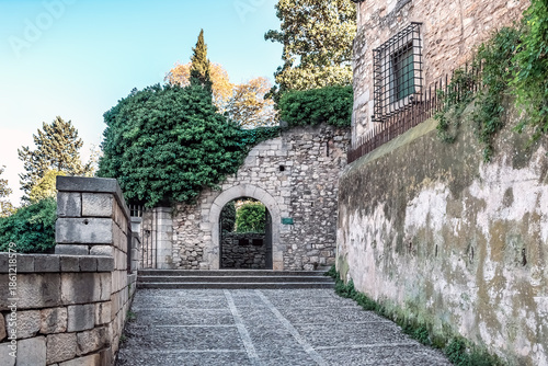 Ancient stone archway entrance to Jardins de la Francesa in Girona, Spain, framed by ivy and greenery. A cobblestone path leads through the medieval passage to a hidden garden