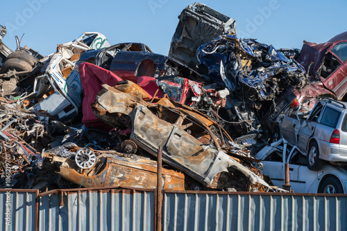 Fotografie a massive heap of crushed and mangled vehicles at a scrap yard, featuring a burnt-out car chassis in the foreground behind a corrugated metal fence