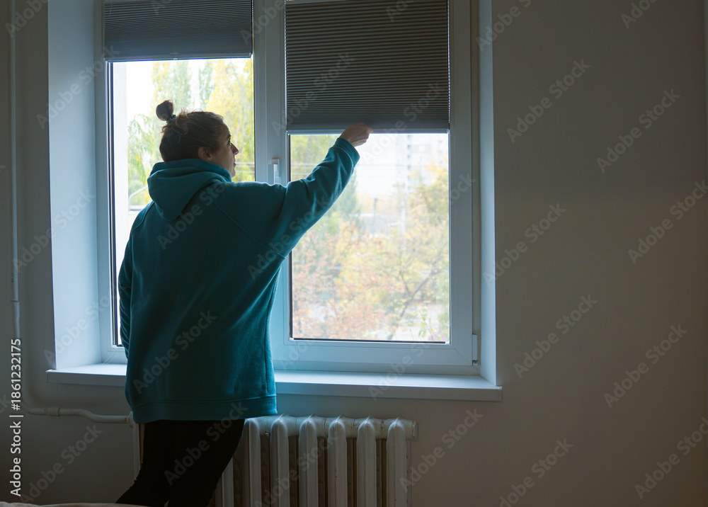 Naklejka premium A woman in a sweatshirt raises the blinds on a large window in a house with natural lighting and a view of the street.
