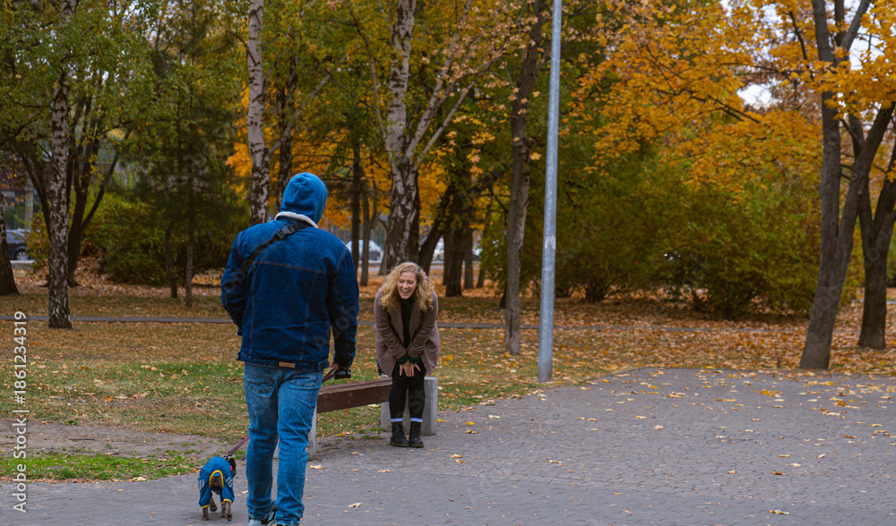Fototapeta premium A man in a blue hoodie walks his dog past a laughing woman in a park filled with autumn leaves.