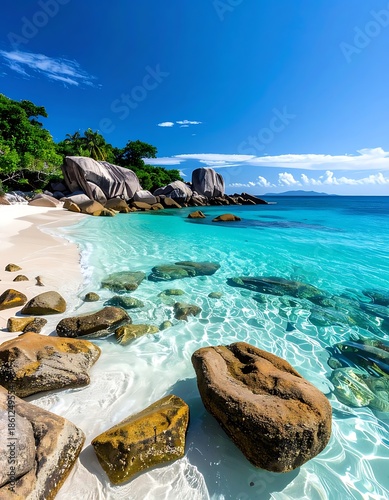 Tropical beach with turquoise water and granite rocks meets lush green vegetation under clear blue sky and scattered clouds