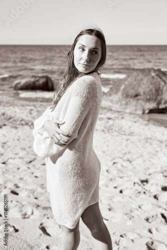 Young woman in soft sweater stands on Baltic Sea sandy shore with rocky background