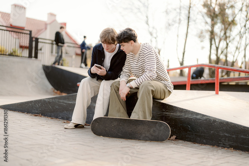 Two young friends sit closely together on a ramp at the skate park. One leans in, focused on his phone, while the other smiles, enjoying the moment in the warm afternoon sun.