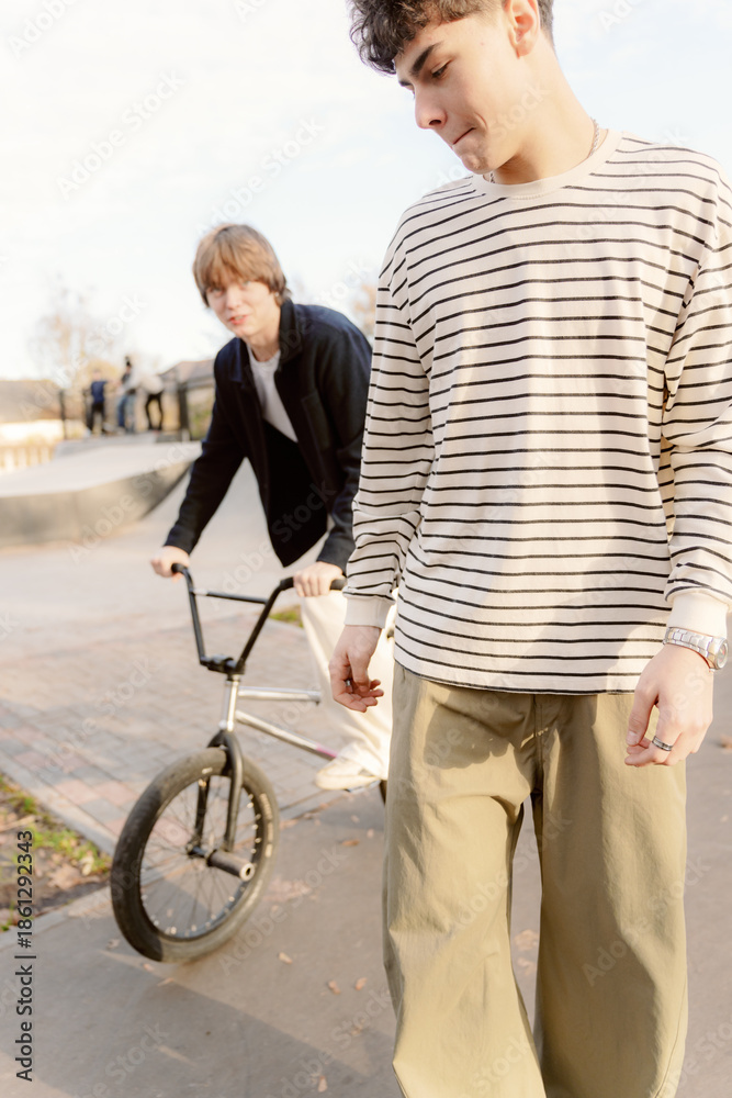 Naklejka premium Two friends take in the warm sun at the skatepark. One rides a BMX bike, while the other observes closely, both showing a relaxed vibe in stylish outfits. It’s a perfect moment of friendship.