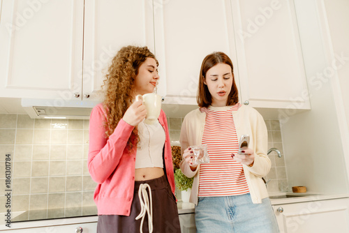 Two young women stand together in a sunny kitchen. One holds a cup, while the other checks her phone. They share a moment of laughter and warmth, enjoying their time together.