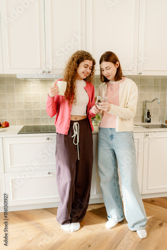 Two friends stand together in a modern kitchen, sipping drinks and sharing laughter. The warm sunlight fills the space, highlighting joyful moments and a sense of togetherness.