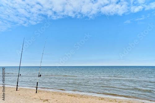 A calm sandy beach with fishing rods set along the shoreline by the quiet sea. A clear blue sky and gentle waves.