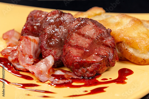 Grilled filet mignon with bacon and baked potato served with a red sauce on a yellow plate