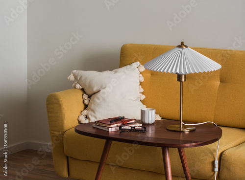 The interior of the living room - a yellow sofa, a wooden table with a lamp, a glass of coffee, and a notebook. Minimalistic interior style