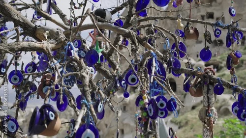 Several nazar boncuk, traditional blue turkish evil eye charms, are suspended from the tree branches as protective talismans, set against the picturesque backdrop of the rocky cappadocia landscape