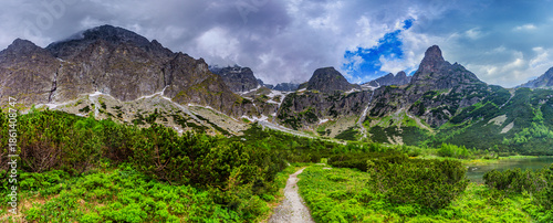 A dramatic landscape shot of a hiking path at the edge of Green Lake (Zelene pleso). Massive granite peaks with snow patches are shrouded in heavy grey storm clouds. Tatra National Park, Slovakia.
