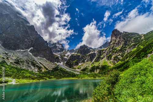 A stunning wide panorama of Zelene pleso (Green Lake) with the historic mountain hut and jagged granite peaks reflecting in the crystal clear turquoise water. High Tatras National Park, Slovakia.