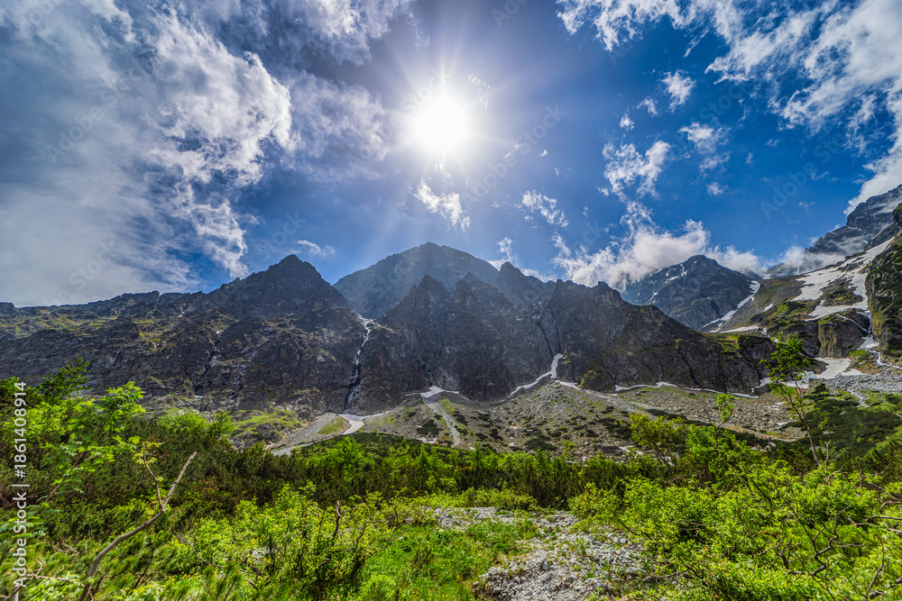 Obraz premium A powerful mountain landscape featuring high granite walls with snow patches under a bright sun with rays. Lush green bushes and rocky terrain in the foreground, located in the Tatra National Park, Sl