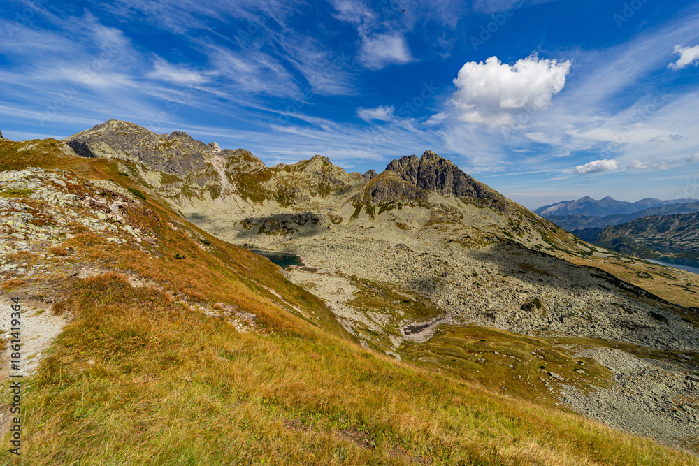 Fototapeta premium A narrow rocky path winds up a steep grassy slope toward a mountain summit. Scenic view from Hladke sedlo in High Tatras National Park, featuring a turquoise lake in the valley under a cloudy blue sky