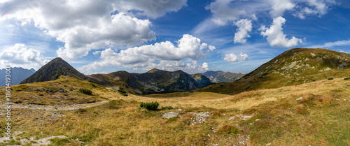 Wide panoramic view of a mountain ridge and vast valleys in the High Tatras National Park. A scenic hiking path leads through grassy slopes near Hladke sedlo under a blue sky with fluffy clouds.