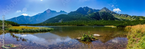 A stunning wide panorama of the crystal clear Velke Biele pleso mountain lake. Surrounded by majestic peaks like Jastrabia veža and lush green slopes under a clear blue sky in the Tatra National Park.
