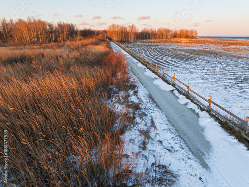 High angle drone view of a snowy path and golden reed fields at Paljassaare peninsula in Tallinn, Estonia during sunset.