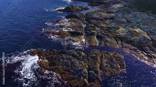 Rough stones rise from the deep blue waters as gentle waves lap against their surfaces. A bright sun illuminates the coastline, creating a peaceful, natural scene.