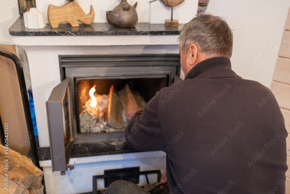 Fototapeta premium Senior male loading wooden log into stove, lights fireplace inside living. Space winter warmth concept.