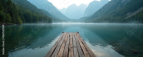 Wooden pier juts into calm lake water with misty forest slopes and distant mountains. Sunrise or sunset paints a peaceful natural landscape ideal for travel inspiration and serene retreats.