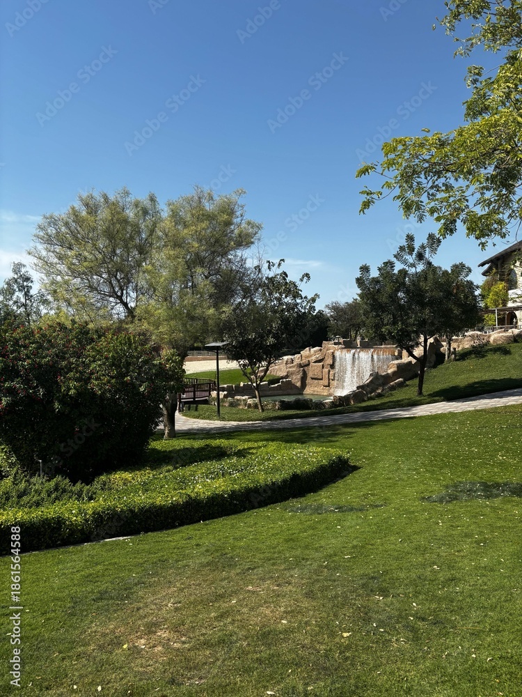 Fototapeta premium A serene park landscape with a small waterfall, lush green grass, trees, and a bright blue sky on a sunny day.