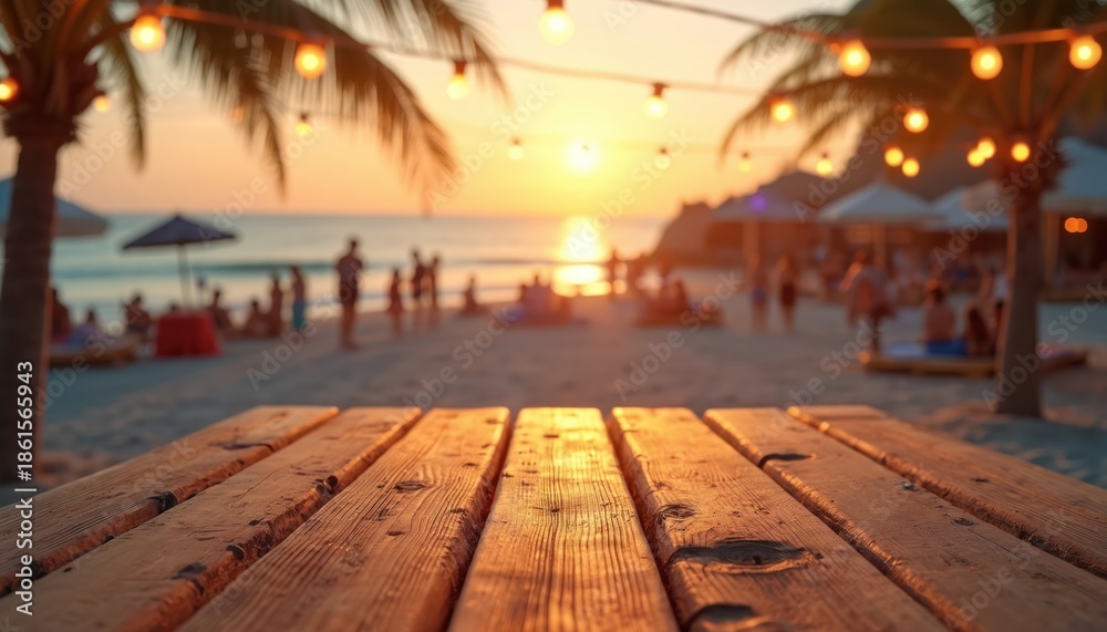 Fototapeta premium Empty wood table foreground. Blurred background beach party scene at sunset. Palm trees and string lights visible. People relax on sand by ocean.