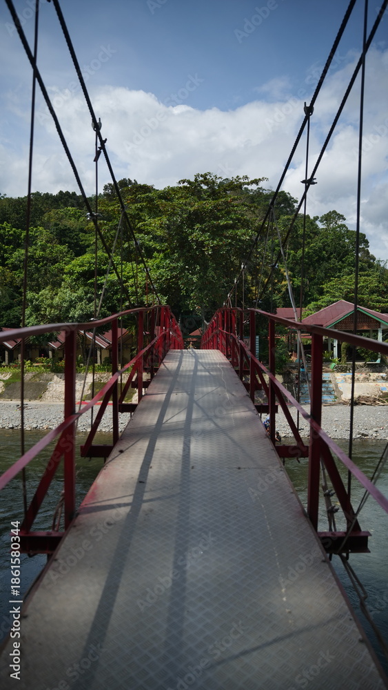 Fototapeta premium Red Metal Suspension Bridge Crossing a River Toward Tropical Village and Forest at Bukit Lawang