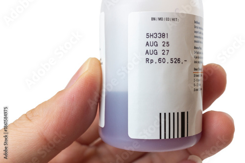 Male hand holding a white plastic medicine bottle with a purple liquid syrup inside. The label clearly shows the manufacturing and expiration dates (AUG 25 and AUG 27), isolated on a white background.