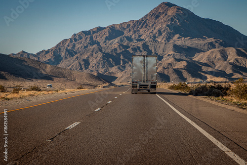 фотографія Following behind a silver 18 wheel truck on a two lane highway in the desert with a rocky hill in the distance