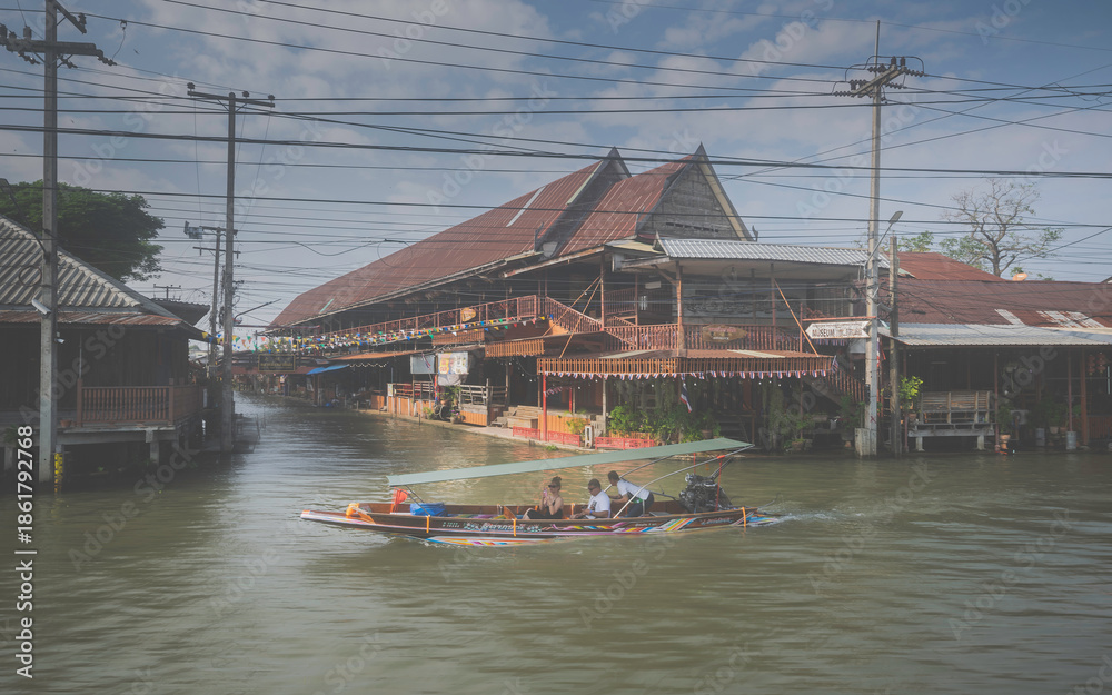 Fototapeta premium A traditional long-tail boat with tourists cruising past a large historic wooden house in a Thai floating market community.