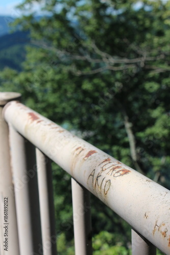 錆びた白い手すりに刻まれた二人の名前と緑の木々のボケ背景｜時間の経過を感じさせる風景と静かな屋外空間