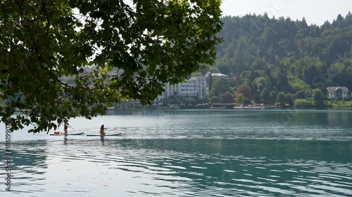 Bled, Slovenia - August 11, 2025: Two people paddleboard on the serene, blue waters of Lake Bled, Slovenia, with lush forested hills and elegant lakeside hotels in the background.