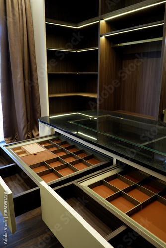 Luxury walk-in closet featuring a central island with a black granite top and glass display. Open drawers show organized jewelry compartments lined with brown leather and walnut wood finish.