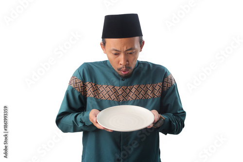 Surprised Asian Muslim man wearing traditional Indonesian Muslim clothing and black peci cap (songkok), looking with surprise at an empty white plate in his hand, Isolated on a white background