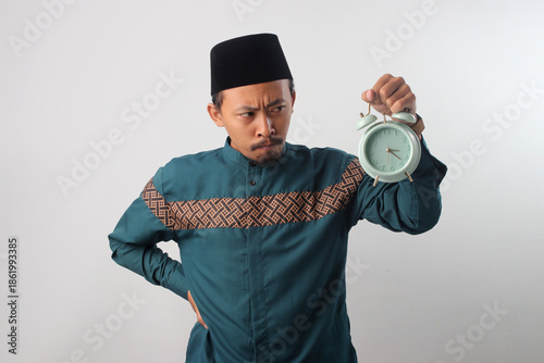Upset Asian Muslim man in a green traditional outfit frowning while looking at an alarm clock in his hand. Isolated on a white background.