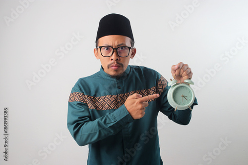 Upset Asian Muslim man in a green traditional outfit pointing at an alarm clock in his hand. Isolated on a white background. Concept of time pressure, lateness, frustration, or deadline stress.