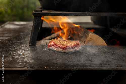 Photos A seasoned steak searing on a hot steel plate over an open wood fire, with flames and smoke creating a rustic outdoor cooking atmosphere focused on heat, texture, and flavor