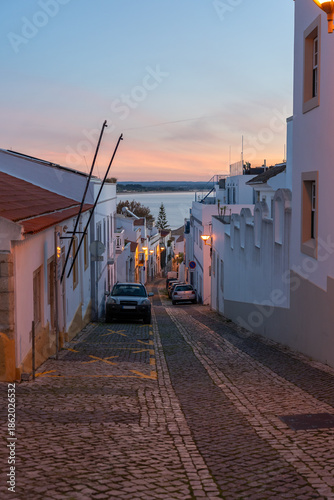 A scenic downhill view of a traditional Portuguese street in Lagos at sunset. The warm glow of street lamps illuminates whitewashed houses, leading the eye toward the calm blue waters of the Atlantic.