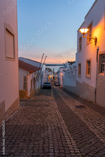 A scenic downhill view of a traditional Portuguese street in Lagos at sunset. The warm glow of street lamps illuminates whitewashed houses, leading the eye toward the calm blue waters of the Atlantic.