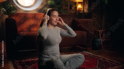 Young woman sitting on a rug at home, meditating with eyes closed and wireless headphones, enjoying calm music, mindfulness, relaxation, and mental wellness in cozy interior