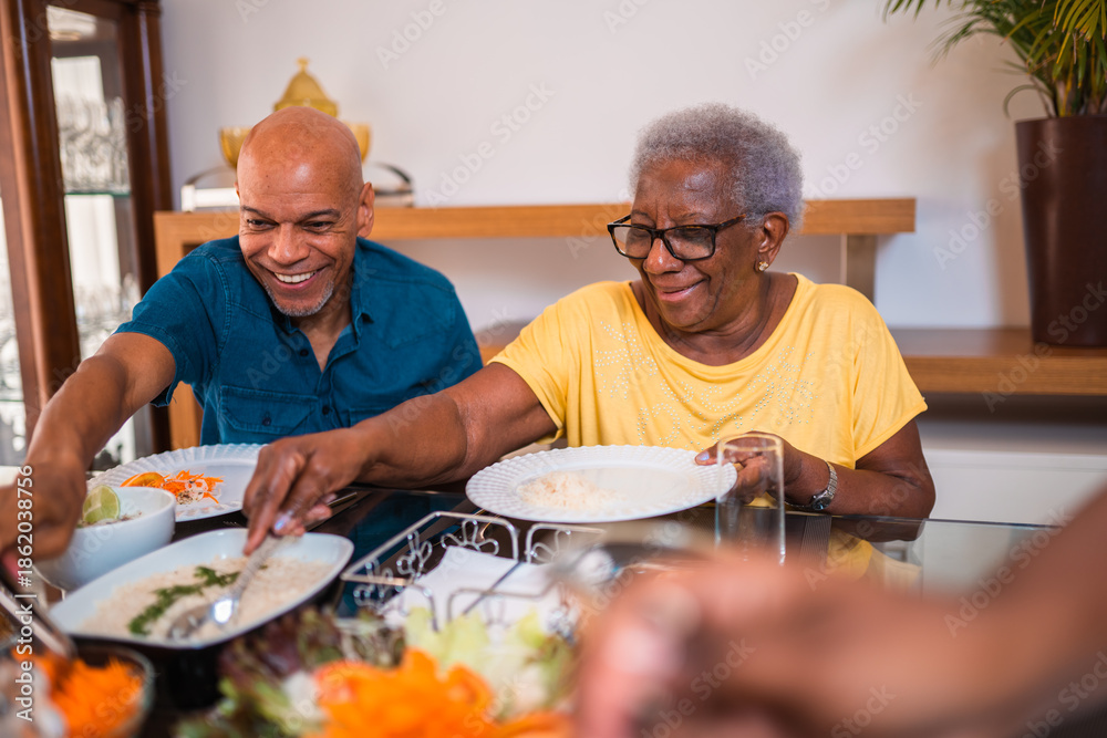 © Nexa - Happy elderly Black woman and adult son sharing a healthy meal together at home, smiling and serving food during a family lunch. © Nexa - Happy elderly Black woman and adult son sharing a healthy meal together at home, smiling and serving food during a family lunch.