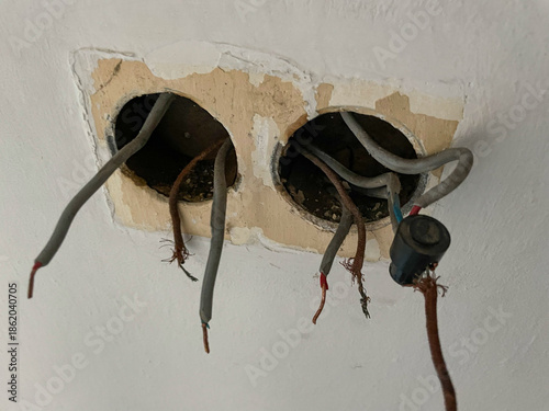 CLOSE UP: Wall cutout reveals exposed electrical wiring with frayed ends, disconnected from their sockets. Surrounding plaster is chipped and damaged, revealing an older electrical system in disrepair
