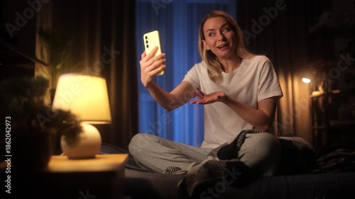 Young woman having a video call on smartphone while sitting on bed at night. Cozy home interior, warm lamp light, casual lifestyle, remote communication, modern technology and online conversation