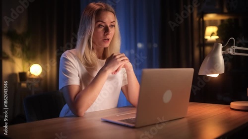 Smiling woman having a video conference on laptop at home at night. Remote work, online meeting, communication, modern home office, warm lighting, casual lifestyle
