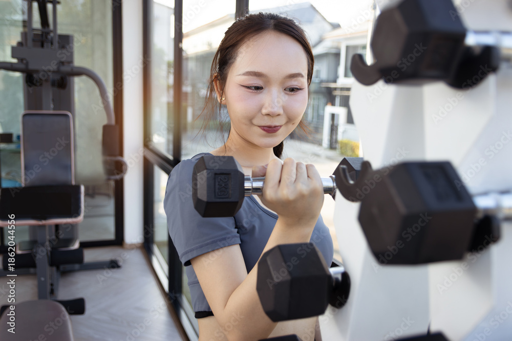 Fototapeta premium Asian smiling beautiful woman lifts dumbbell from dumbbell rack to exercise in sport gym. Young fitness girl engaging in workout exercises, training to keep body strong and fit, healthy bodybuilding.