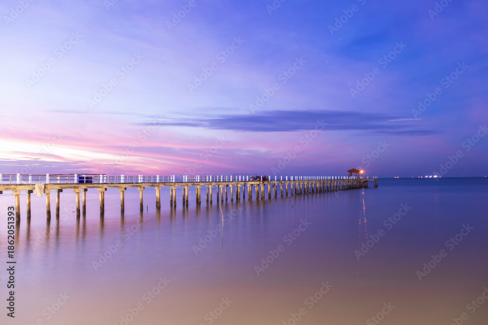 Obraz premium Long wooden pier at dusk with soft purple sky and calm water reflecting lights at Bang Pu in Samut Prakan, Thailand.