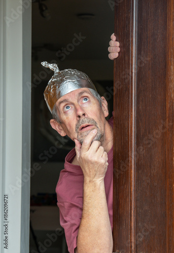 Tinfoil hat picture. A man wearing a tinfoil hat looks anxiously out of a door towards the sky. Paranoia and conspiracy theory. Alien abduction and government cover up. Mind control and brainwashing.