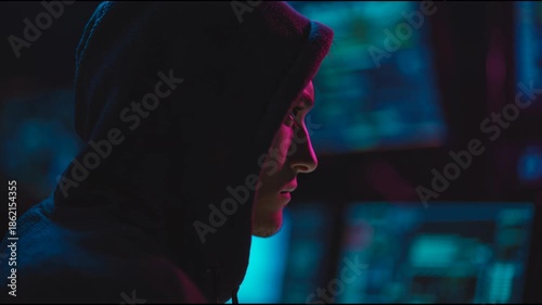 Silhouette of a mysterious hacker in a hoodie working in a dark room with blue screens for cybersecurity and coding concept.