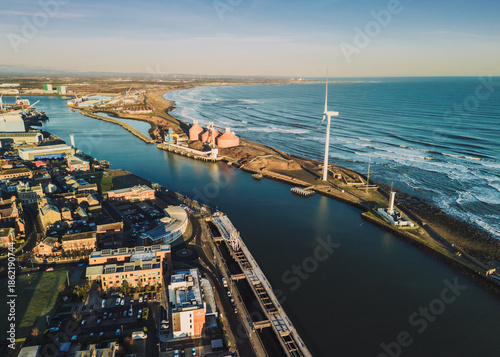 Blyth Northumberland UK: 31st Dec 2025: Drone aerial view of Port of Blyth with docks and industrial buildings along the water. Waves crash along the shore, and a wind turbine stands tall
